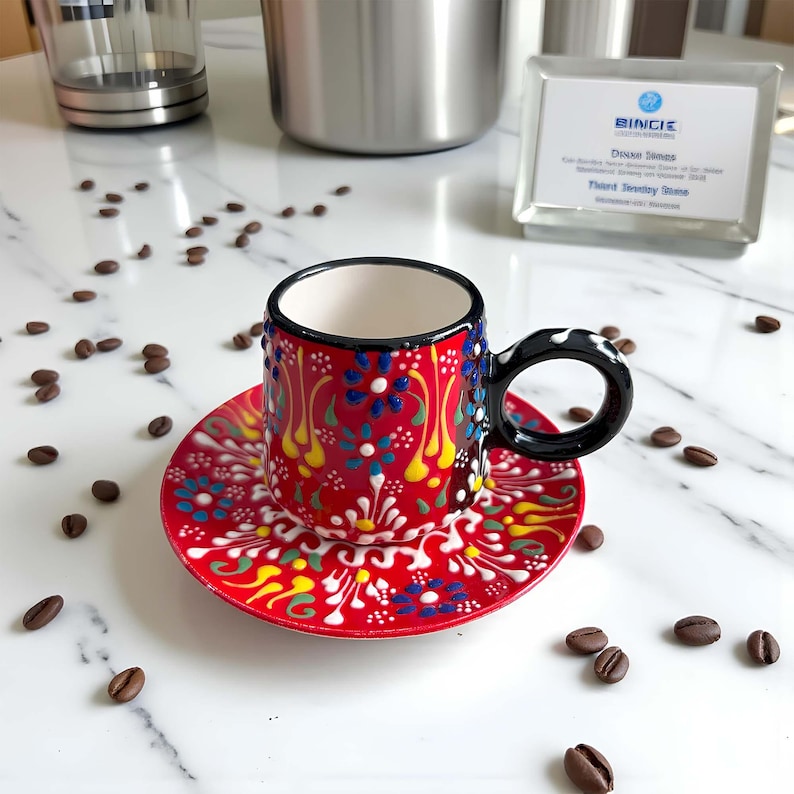 May include: A small, hand-painted ceramic coffee cup and saucer set. The cup and saucer are red with a floral design in blue, yellow, and white. The cup has a black handle with white polka dots. Coffee beans are scattered around the set.