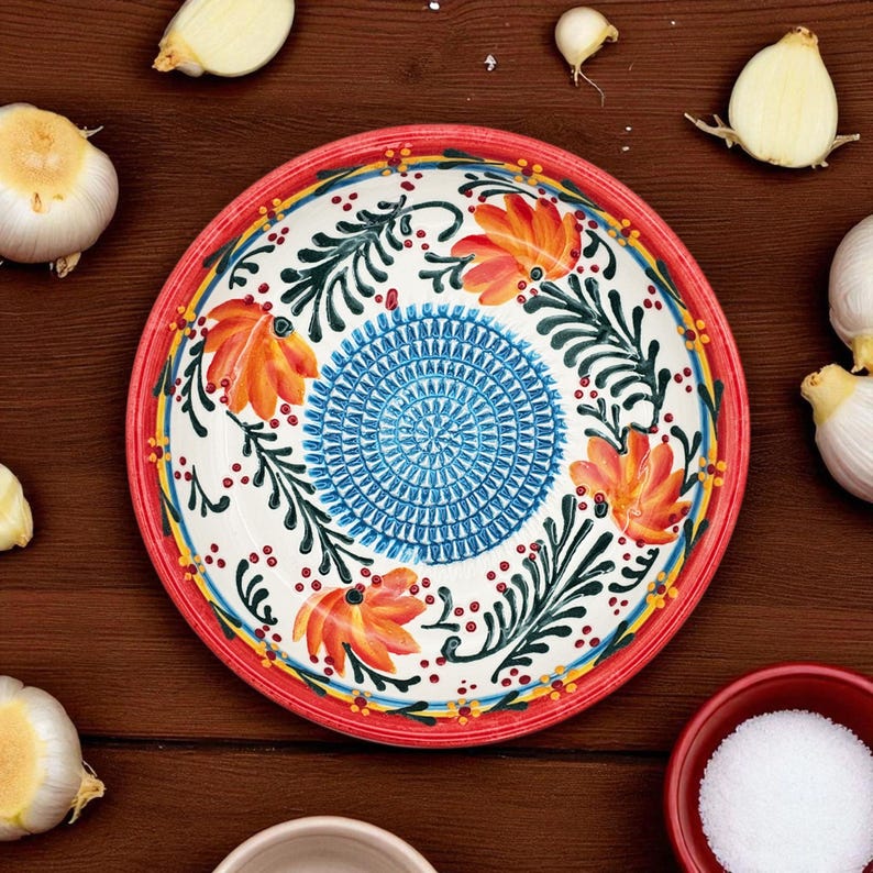 May include: A decorative ceramic plate with a red rim, featuring a floral design with orange flowers, green leaves, and a blue center. Garlic cloves and salt are scattered around the plate on a wooden surface.