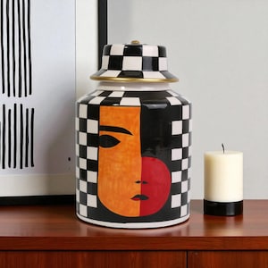 May include: A decorative ceramic jar with a lid, featuring a black and white checkerboard pattern and a painted face design in orange and red. The jar has a gold rim and is next to a white candle.