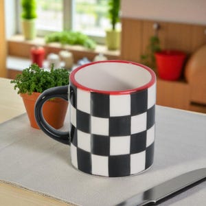Puede incluir: Una taza de cerámica con un diseño a cuadros en blanco y negro. El borde es rojo y el asa es negra. La taza está sobre una superficie gris claro, con una pequeña planta en maceta y un cuchillo en el fondo. La taza está diseñada para bebidas.