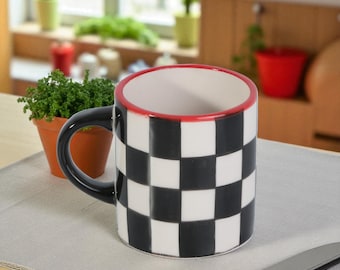 May include: A ceramic mug with a black and white checkered pattern. The rim is red, and the handle is black. The mug sits on a light gray surface, with a small potted plant and a knife in the background. The mug is designed for beverages.