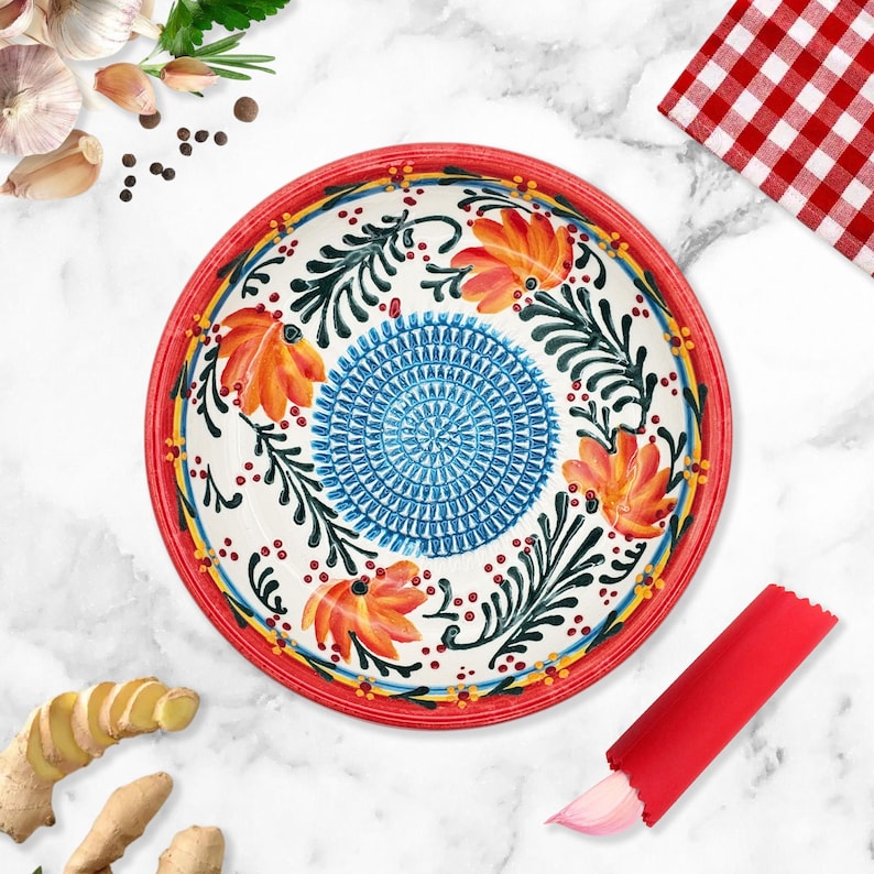 May include: A round, hand-painted ceramic bowl with a red rim and floral design. The bowl features orange flowers, blue and green foliage, and a blue center pattern. A red garlic peeler is next to the bowl.