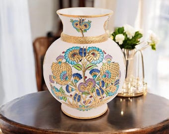 Può includere: Un vaso in ceramica color crema con una base ampia e un collo stretto. Il vaso è adornato da una fascia dorata e da intricati motivi floreali blu, verde acqua, viola e oro. Il vaso è esposto su un tavolo di legno scuro.
