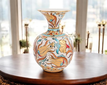 Può includere: Un vaso decorativo in ceramica con una base ampia e una parte superiore svasata. Il vaso presenta uno sfondo bianco con un motivo floreale in oro, arancione, rosso e blu. Il vaso è appoggiato su un tavolo rotondo di legno.