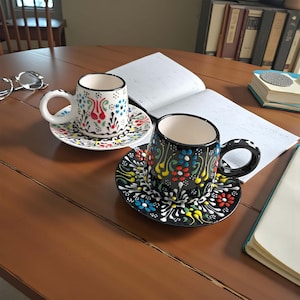 May include: Two decorative ceramic coffee cups with matching saucers. One cup is white with colorful floral designs, and the other is black with similar patterns. Both cups have a curved handle and sit on a wooden table.