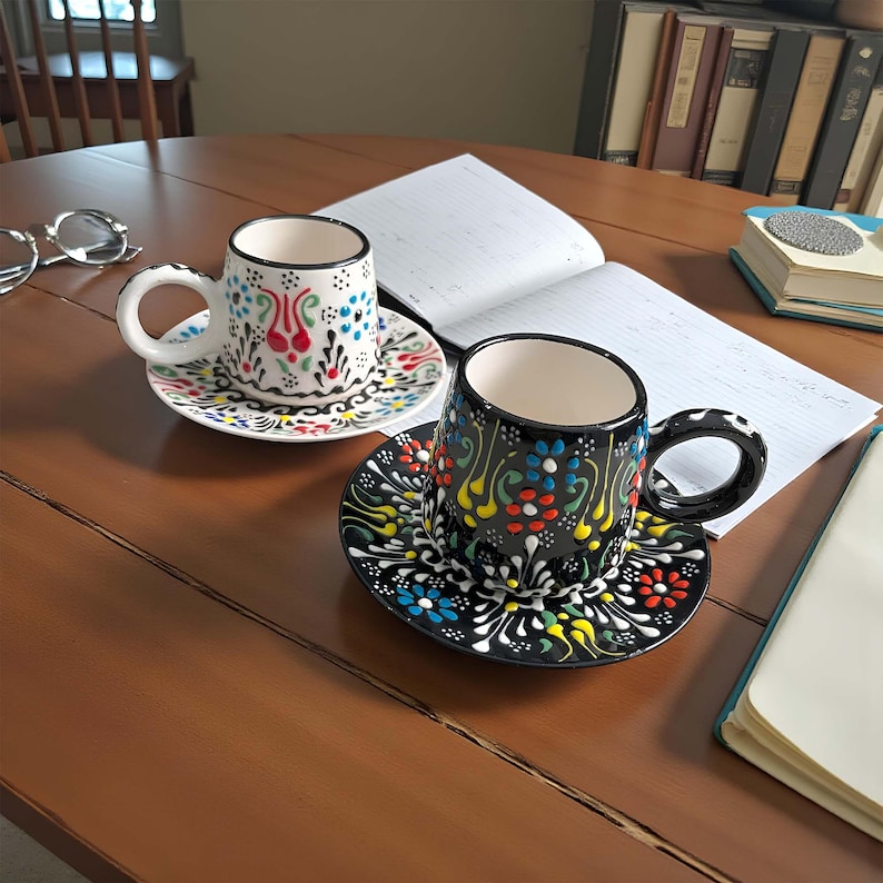 May include: Two decorative ceramic coffee cups with matching saucers. One cup is white with colorful floral designs, and the other is black with similar patterns. Both cups have a curved handle and sit on a wooden table.