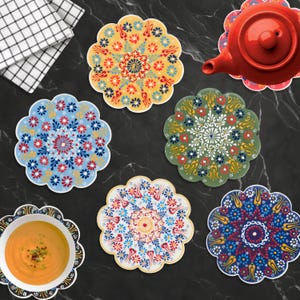 May include: A collection of colorful, floral-patterned ceramic coasters. The coasters feature intricate designs in shades of blue, red, yellow, and green. A red teapot and a bowl of soup are also visible, suggesting a kitchen or dining setting.