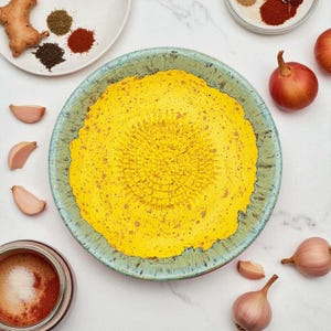 Puede incluir: Un rallador amarillo con una superficie texturizada se encuentra sobre un plato con borde turquesa y verde. Los dientes de ajo, las chalotas y las especias están dispuestas alrededor del plato. Jengibre y otras especias están en un plato blanco. También se ve un tarro de especias.