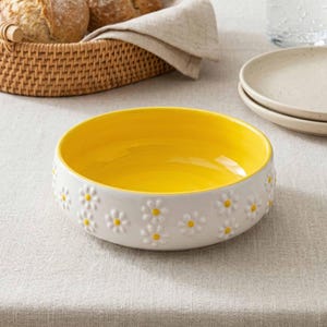 May include: A white ceramic bowl with a bright yellow interior and daisy flower detailing around the exterior. The bowl is on a neutral-coloured tablecloth, with a wicker basket of bread and plates in the background.