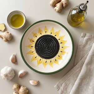Puede incluir: Un plato rallador de cerámica con forma de girasol, con borde verde, pétalos amarillos y centro negro. El plato está rodeado de ingredientes como ajo, jengibre y aceite de oliva, lo que sugiere un tema culinario. También hay una botella de aceite de oliva y un paño de lino.