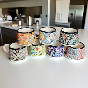 May include: A selection of ceramic mugs featuring vibrant floral patterns. Each mug has a black rim and a white interior. The designs showcase orange, blue, green, and red flowers. One mug includes a handle. The mugs are displayed on a white surface.