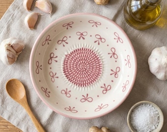 Puede incluir: Un rallador de ajo de cerámica blanca con borde rosa y diseño de lazo. El centro tiene una superficie ralladora texturizada. Ajos, una cuchara de madera, jengibre, aceite de oliva y sal están dispuestos alrededor del rallador.