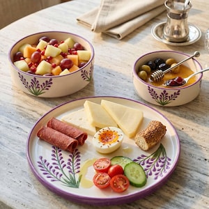 Puede incluir: Un desayuno con un plato y cuencos blancos con un diseño floral morado. El plato contiene queso en rodajas, carne enrollada, un huevo pasado por agua, tomates y pepinos. Los cuencos contienen fruta, aceitunas y miel. También hay un vaso y una servilleta.