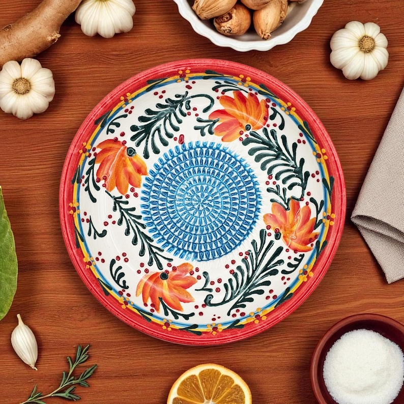 May include: A decorative ceramic plate with a red rim, featuring a floral design with orange flowers, green leaves, and a blue center. The plate is surrounded by ingredients like garlic, nuts, and a lemon slice, suggesting a culinary theme.