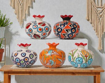 May include: Five decorative ceramic pomegranate vases with red, orange, and black accents. The vases feature intricate patterns of circles, flowers, and geometric designs. They are displayed on a wooden table and a white shelf.