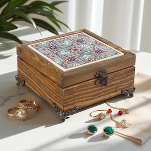 May include: A wooden jewelry box with a decorative tile top featuring a floral pattern in red, blue, and white. The box has metal corner feet and a clasp. Several pieces of jewelry, including rings and earrings, are displayed nearby.