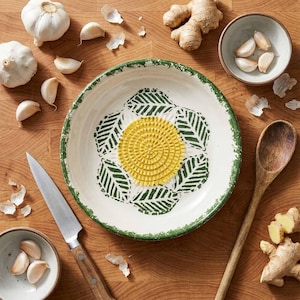 Puede incluir: Un cuenco rallador de ajo de cerámica blanca con borde verde y diseño de hojas. Un centro texturizado amarillo está rodeado de hojas verdes. Ajos, jengibre, una cuchara de madera y un cuchillo están dispuestos sobre una superficie de madera.