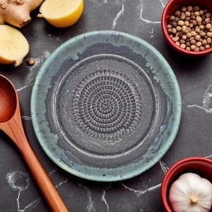 Puede incluir: Un plato rallador de cerámica con un centro gris oscuro y un borde verde azulado. El plato tiene una superficie texturizada con un patrón circular en el centro. Ingredientes como ajo, granos de pimienta, jengibre y limón están dispuestos alrededor del plato.