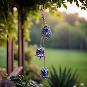 May include: Three blue and white ceramic wind chimes with a floral design hang from a natural rope. The chimes are suspended from a tree branch, with colourful beads and a blurred green background.
