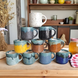 May include: A selection of handmade ceramic mugs in various colours, including speckled white, black, yellow, brown, blue, and teal. Each mug features a unique glaze design and a sturdy handle. The mugs are stacked on a wooden surface.