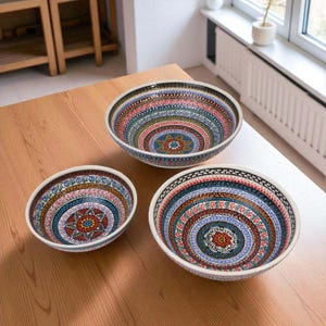 May include: Three decorative ceramic bowls with intricate, colorful patterns. The bowls feature a white rim and a detailed design in shades of red, blue, and gold. The bowls are arranged on a wooden table, showcasing their varying sizes.