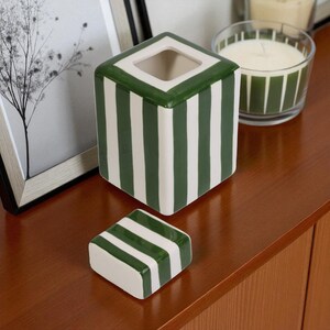 Pu&ograve; includere: Un contenitore quadrato in ceramica con coperchio rimovibile. Il contenitore e il coperchio sono bianchi con strisce verdi verticali. Il bordo superiore del contenitore &egrave; verde. Una candela in un barattolo di vetro &egrave; sullo sfondo.