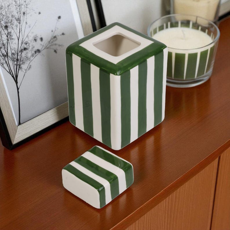 Pu&ograve; includere: Un contenitore quadrato in ceramica con coperchio rimovibile. Il contenitore e il coperchio sono bianchi con strisce verdi verticali. Il bordo superiore del contenitore &egrave; verde. Una candela in un barattolo di vetro &egrave; sullo sfondo.