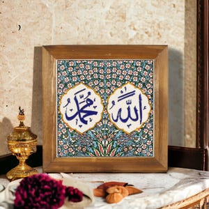 Può includere: Piastrella quadrata con cornice in legno con motivo floreale e calligrafia araba blu. La piastrella presenta i nomi "Muhammad" e "Allah" in elegante scrittura. La cornice è di colore marrone chiaro.
