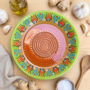 Puede incluir: Un plato rallador de cerámica con borde verde y centro marrón. El borde presenta un diseño floral en rojo, azul y blanco. También se ven ajo, jengibre, aceite de oliva y una cuchara de madera.