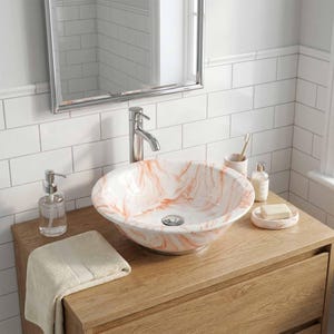 May include: A white and orange marble-patterned vessel sink sits atop a wooden vanity. A chrome faucet, mirror, and soap dispenser are visible. A folded beige towel and soap dish with a bar of soap are also present. The bathroom has white tile walls.