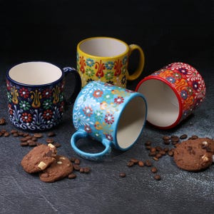 May include: Four ceramic mugs with colorful floral patterns. The mugs are in shades of blue, yellow, and red, with white interiors. Chocolate chip cookies and coffee beans are scattered around the mugs on a dark surface.