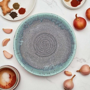 Puede incluir: Un plato rallador de cerámica gris con borde turquesa, rodeado de dientes de ajo, cebollas rojas y pequeños platos de especias. El plato tiene un centro texturizado para rallar ingredientes. Tema culinario.