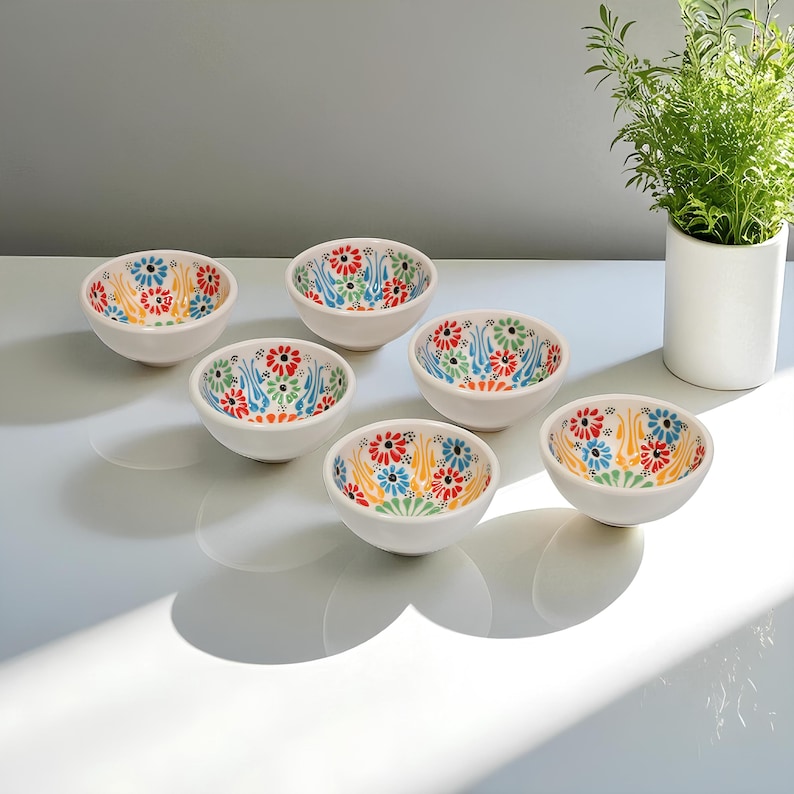 May include: Six small, white ceramic bowls with colorful floral patterns. The interior of each bowl features a vibrant design of red, blue, yellow, and green flowers. The bowls are arranged on a white surface, with a plant in a white pot in the background.