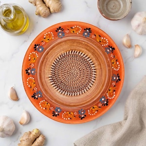 Puede incluir: Plato rallador de cerámica naranja con centro texturizado. El borde presenta diseños florales en azul, amarillo y blanco. A su alrededor, dientes de ajo, jengibre y aceite de oliva.