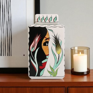 May include: A white ceramic decorative jar with a lid, featuring a painted face and floral designs in red, black, and yellow. A lit candle in a glass holder sits nearby on a wooden surface.