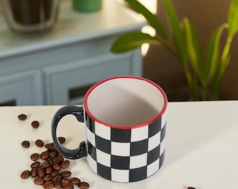 May include: A ceramic coffee mug with a black and white checkered pattern. The rim is red, and the handle is black. Coffee beans are scattered around the mug on a light-colored surface.