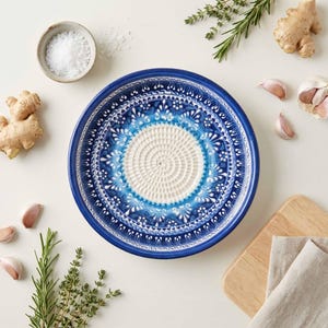May include: A blue and white ceramic garlic grater with a decorative floral pattern. The center has a textured surface for grating. Ingredients like garlic, ginger, salt, and herbs surround the grater.