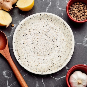 Puede incluir: Un plato de cerámica blanca moteada es el punto focal de esta toma cenital. El plato está rodeado de ingredientes como una cuchara de madera, jengibre, mitades de limón, granos de pimienta en un cuenco rojo y un bulbo de ajo en un cuenco rojo, todo sobre una superficie oscura.