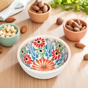 May include: A white ceramic bowl with a colorful floral design in red, blue, green, and orange. The bowl is surrounded by small bowls filled with nuts, including almonds and cashews, on a wooden surface.
