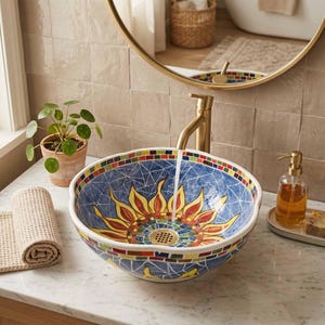 May include: A round ceramic vessel sink featuring a blue mosaic background with a central yellow and red sun design. The rim is decorated with a multi-colored tile pattern. A gold-toned faucet is positioned above the basin, with water flowing.