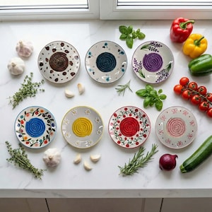 Puede incluir: Una colección de siete platos ralladores de cerámica con diseños coloridos. Cada plato presenta una superficie de rallado texturizada central, rodeada de bordes florales o estampados. Verduras frescas y hierbas aromáticas están dispuestas alrededor de los platos.