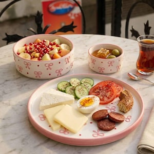 May include: A breakfast arrangement with a white and pink bow-patterned plate and bowls. The plate displays sliced cucumbers, tomatoes, cheese, eggs, and sausage. Bowls contain fruit salad and honey with olives. A glass of tea is also visible.