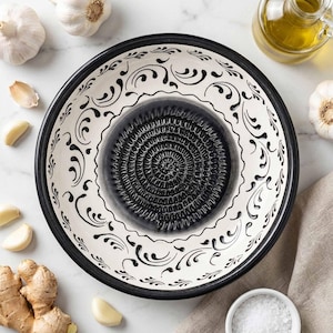 Puede incluir: Un cuenco rallador de ajo de cerámica blanca y negra con un diseño decorativo de remolino. El centro del cuenco tiene una superficie texturizada para rallar. Los dientes de ajo, el jengibre, el aceite de oliva y la sal están dispuestos alrededor del cuenco.