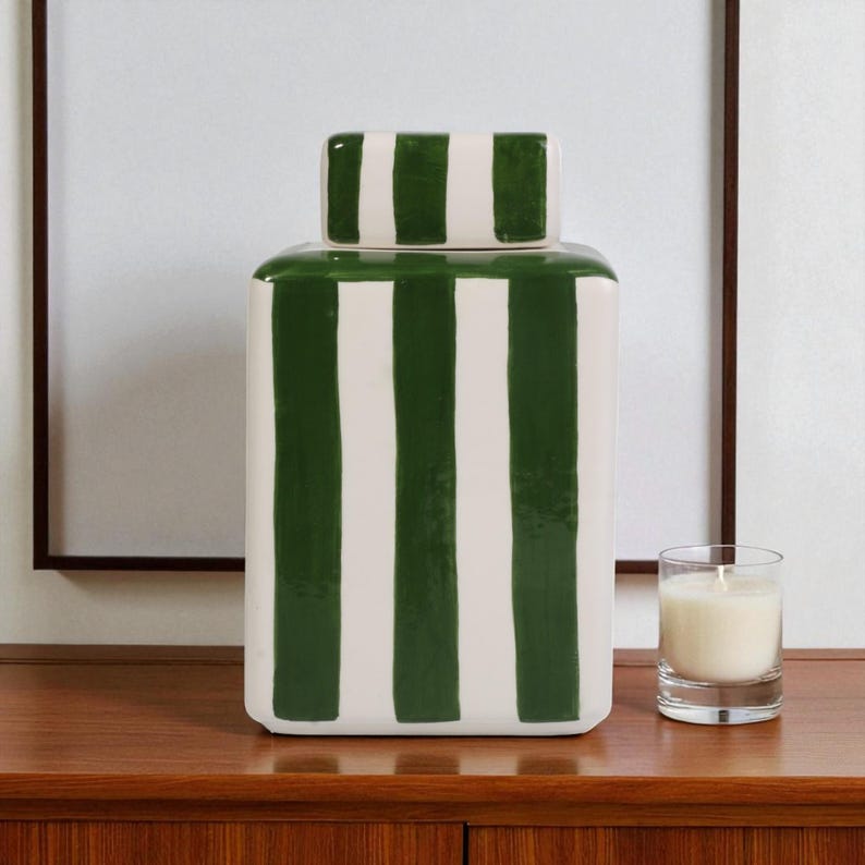 Pu&ograve; includere: Un vaso rettangolare in ceramica con coperchio, decorato con strisce verdi verticali su sfondo bianco. A destra, una piccola candela in un bicchiere trasparente. Il vaso &egrave; su una superficie di legno.