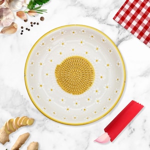 May include: A white ceramic garlic grater with a gold center and rim, decorated with small white daisies. A red garlic peeler and a clove of garlic are next to the grater. The background includes garlic, ginger, and a red and white checkered cloth.