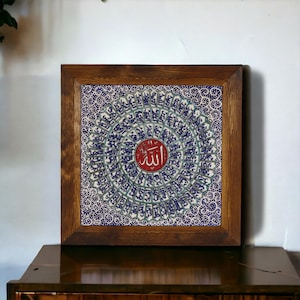 May include: Square framed artwork featuring a detailed blue and white circular pattern with a red central medallion. The artwork is encased in a dark brown wooden frame, creating a striking contrast. The central medallion features Arabic calligraphy.