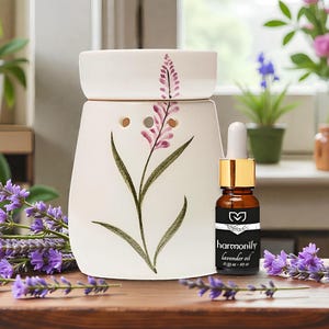 Puede incluir: Un difusor de aceites esenciales de cerámica blanca con un diseño floral de lavanda, junto con una pequeña botella de aceite de lavanda. La botella tiene una tapa dorada y una etiqueta que dice "harmony lavender oil". Ramas de lavanda fresca están dispuestas alrededor del difusor.
