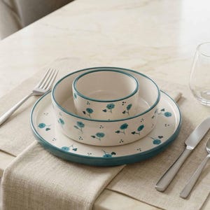 Op de afbeelding: Een crèmekleurige keramische serviesset met een turquoise rand en bloemenpatroon. De set bevat een bord, een kom en een kleinere kom. Bestek en een glas zijn ook zichtbaar op een neutraal gekleurde placemat.