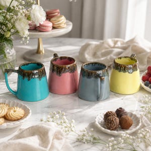 Op de afbeelding: Vier keramische mokken in turquoise, roze, leiblauw en geel, elk met een donkerbruin druipglazuurpatroon rond de rand. De mokken hebben een gladde, glanzende afwerking en een stevig handvat, gerangschikt op een wit marmeren oppervlak.