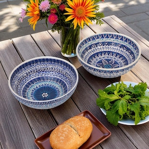 May include: Two blue and white patterned ceramic bowls are displayed on a wooden table, with a plate of green leafy vegetables and a loaf of bread. A vase of colorful flowers is in the background. The bowls have a decorative design.